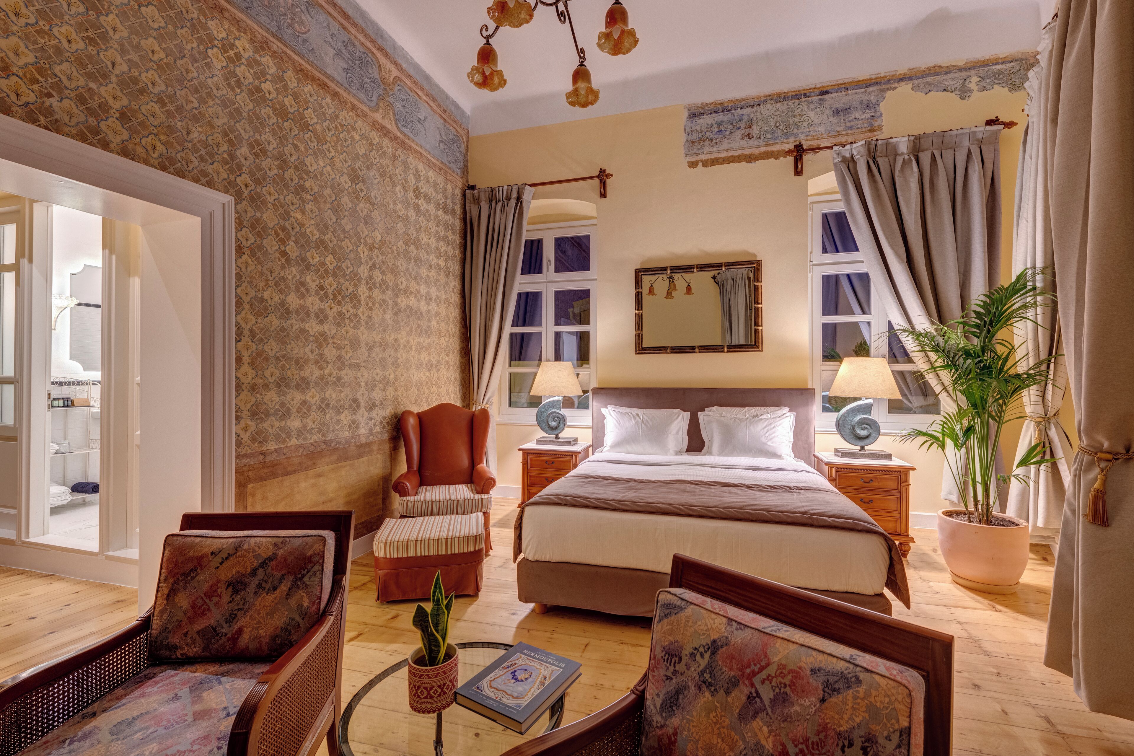 A spacious hotel room with a large bed, two armchairs, a wooden coffee table with a book, a potted plant, and a view of the outside through a window.