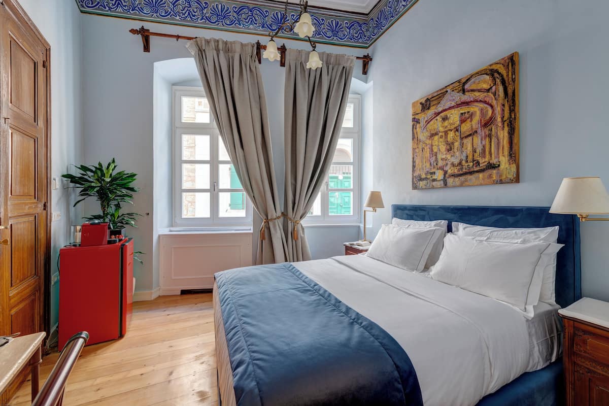 A bedroom with a bed, curtains, a painting, a plant, and a red appliance.