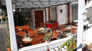 Een terras met houten banken en tafels, plantenbakken en een rode deur.