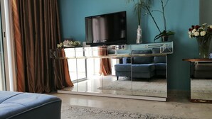 A modern living room with a large flat-screen TV mounted on the wall, a blue sofa, a glass coffee table, and a decorative vase with flowers. - Superbe appartement avec Piscine (La Soukra)