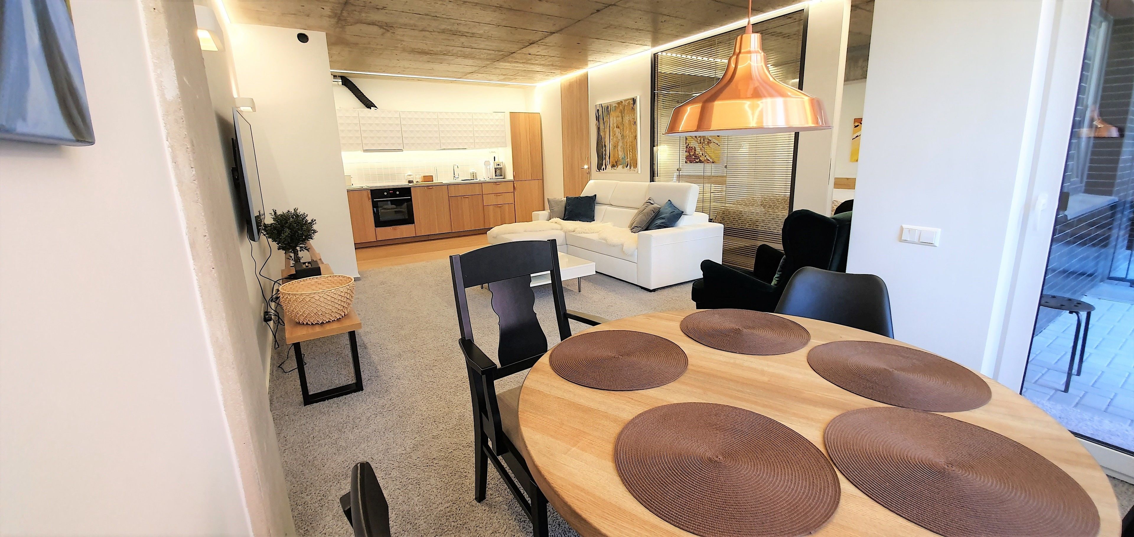 A modern living room with a dining table, chairs, and a sofa. There is a kitchen in the background with wooden cabinets and a large window.