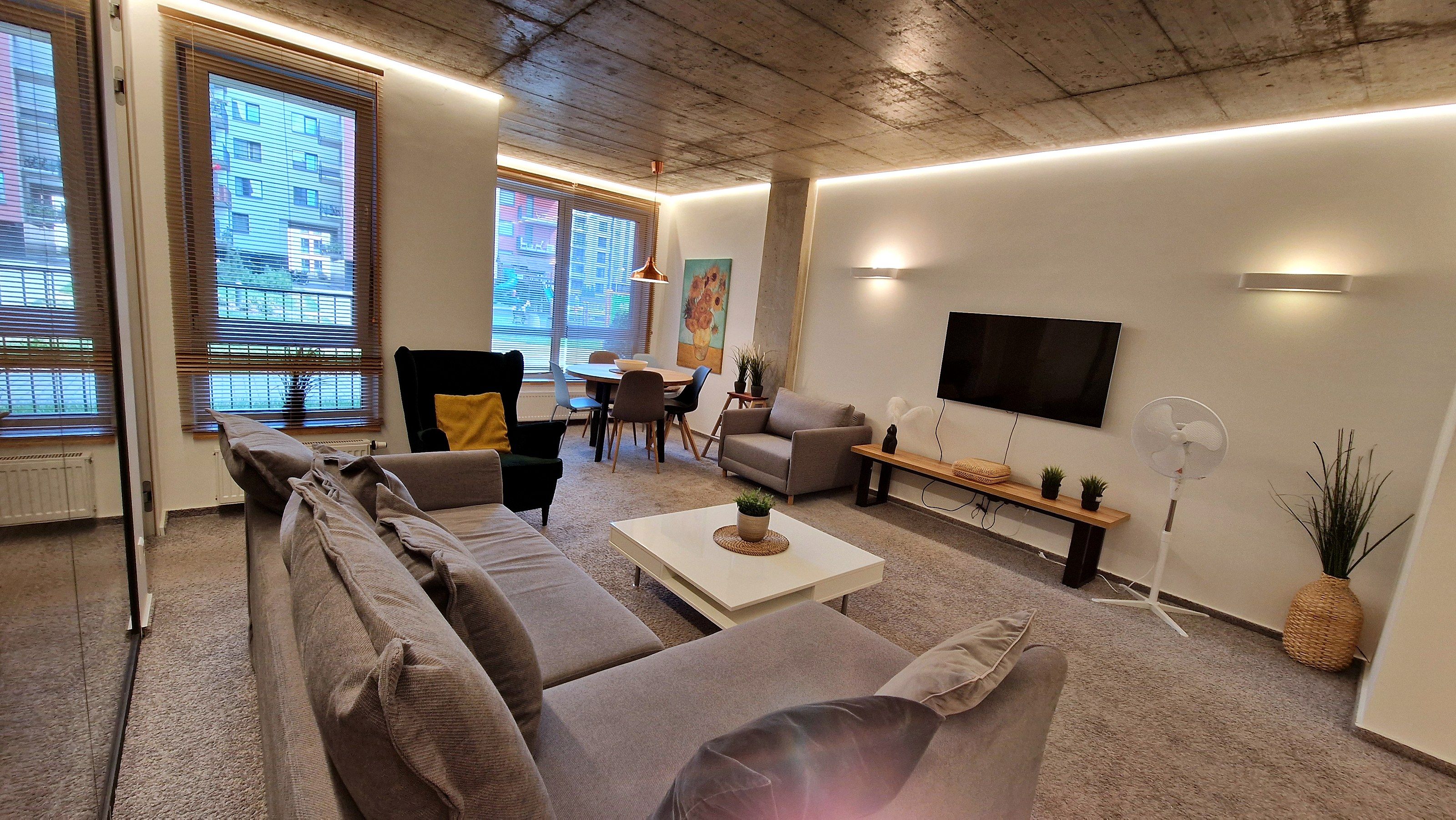 A modern living room with a sofa set, a dining table with chairs, a TV mounted on the wall, and a large window with blinds.