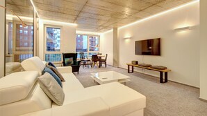 A modern living room with a white sofa, a dining area with chairs and a table, a flat-screen TV mounted on the wall, and large windows offering views of the city. - Kauno-Algirdo Luxury Vilnius Apartments (Vilnius)