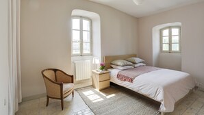 A bedroom with a bed, nightstand, chair, and two windows. - Charming apartment in town house in the heart of Eygalieres (Eygalières)