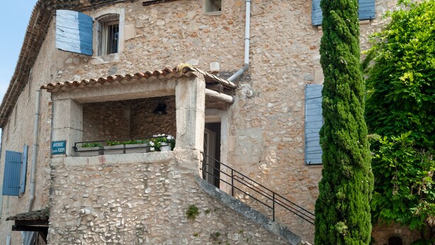 A stone building with a tiled roof, a staircase leading to an entrance, and a tall green tree beside it. - Charming apartment in town house in the heart of Eygalieres (Eygalières)
