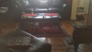 Una habitación con suelo de madera, un tapete estampado, un sofá de cuero negro, una mesita y una ventana con luz natural.