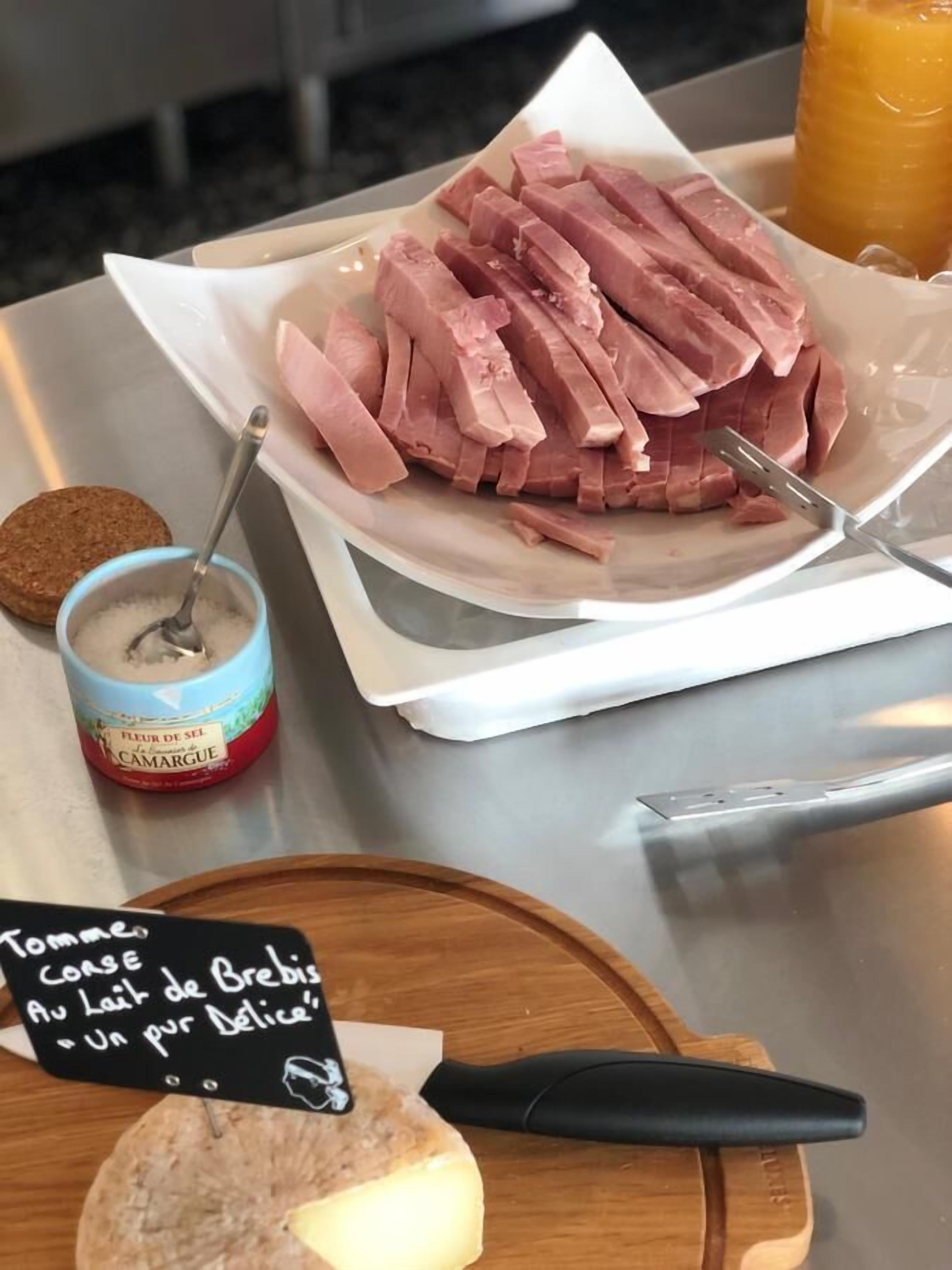 A plate of sliced ham, a small container of jam, and a wooden board with a piece of cheese and a knife.