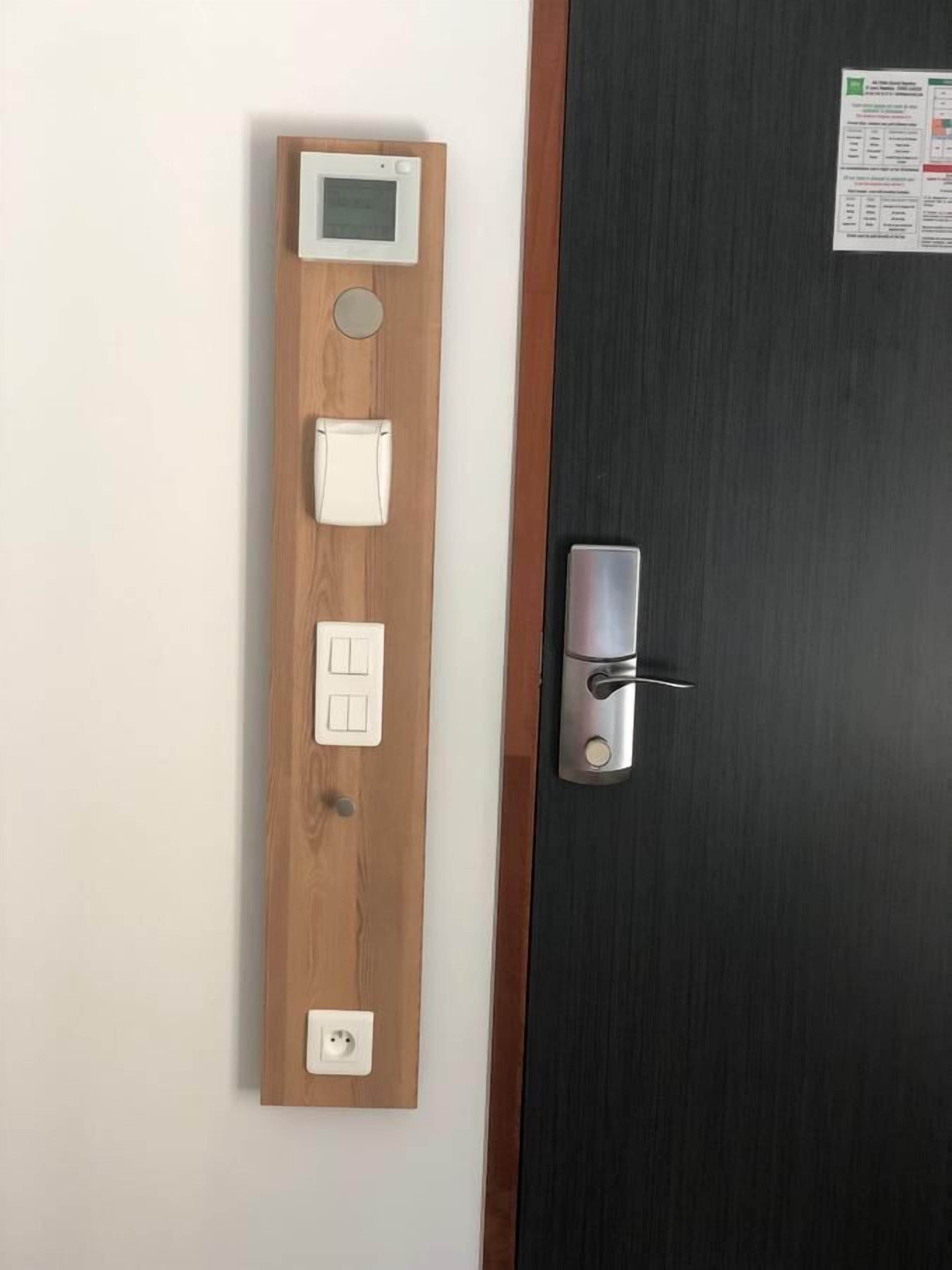 Panel de madera con pantalla digital, dos interruptores y un enchufe, fijado a una pared junto a una puerta con picaporte metálico.