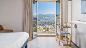 Ett hotellrum med en säng, ett skrivbord och en stol, samt ett stort fönster med utsikt över en småbåtshamn och berg.