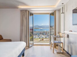 Habitación de hotel con cama, escritorio y silla, un ventanal con vistas a un puerto deportivo y montañas.