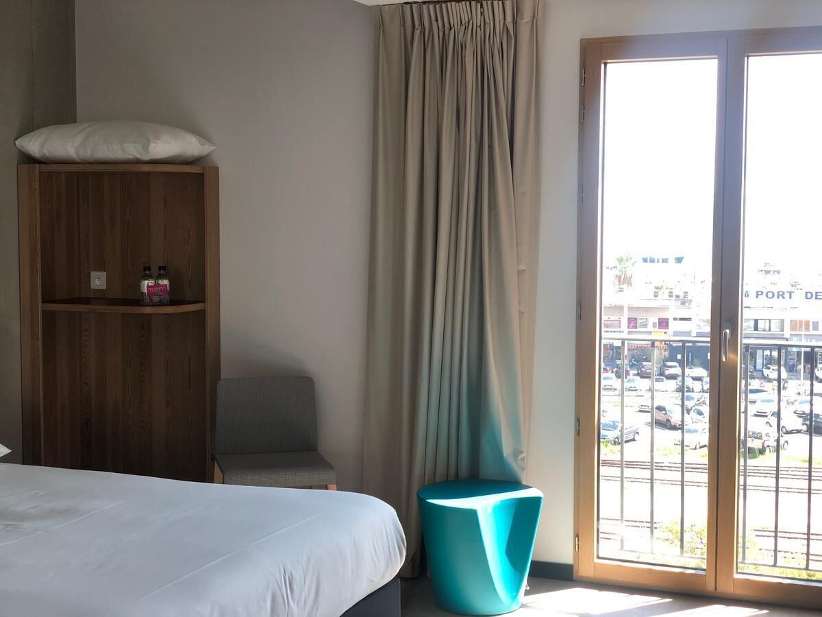 A hotel room with a bed, a wooden wardrobe, a teal chair, a window with a view of a parking lot and buildings.