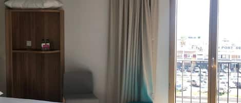 A hotel room with a bed, a wooden wardrobe, a teal chair, a window with a view of a parking lot and buildings.