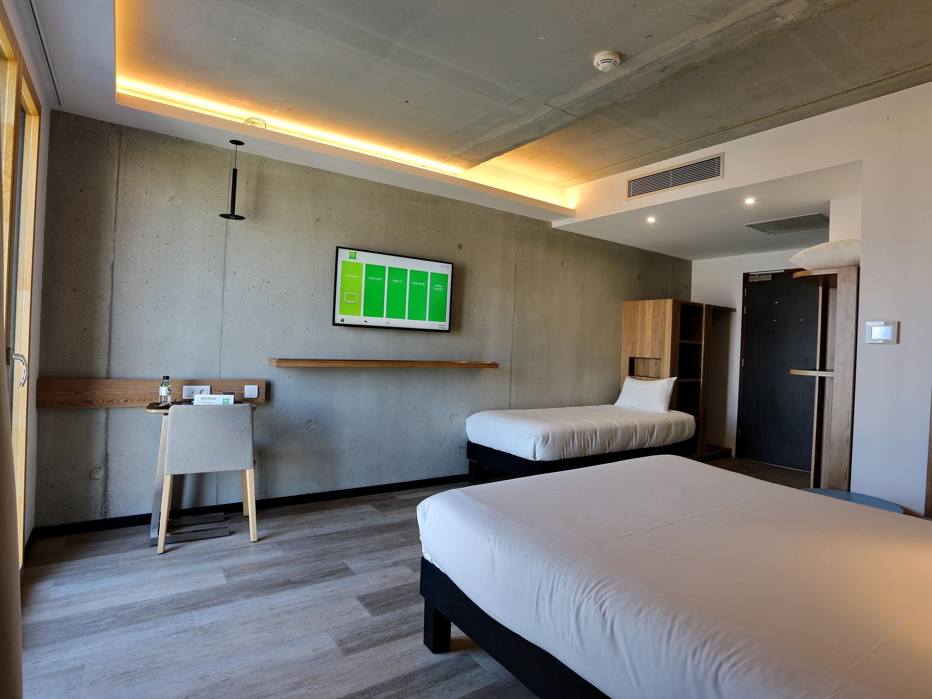 A modern hotel room with a large bed, a small desk, a chair, and a TV mounted on the wall.