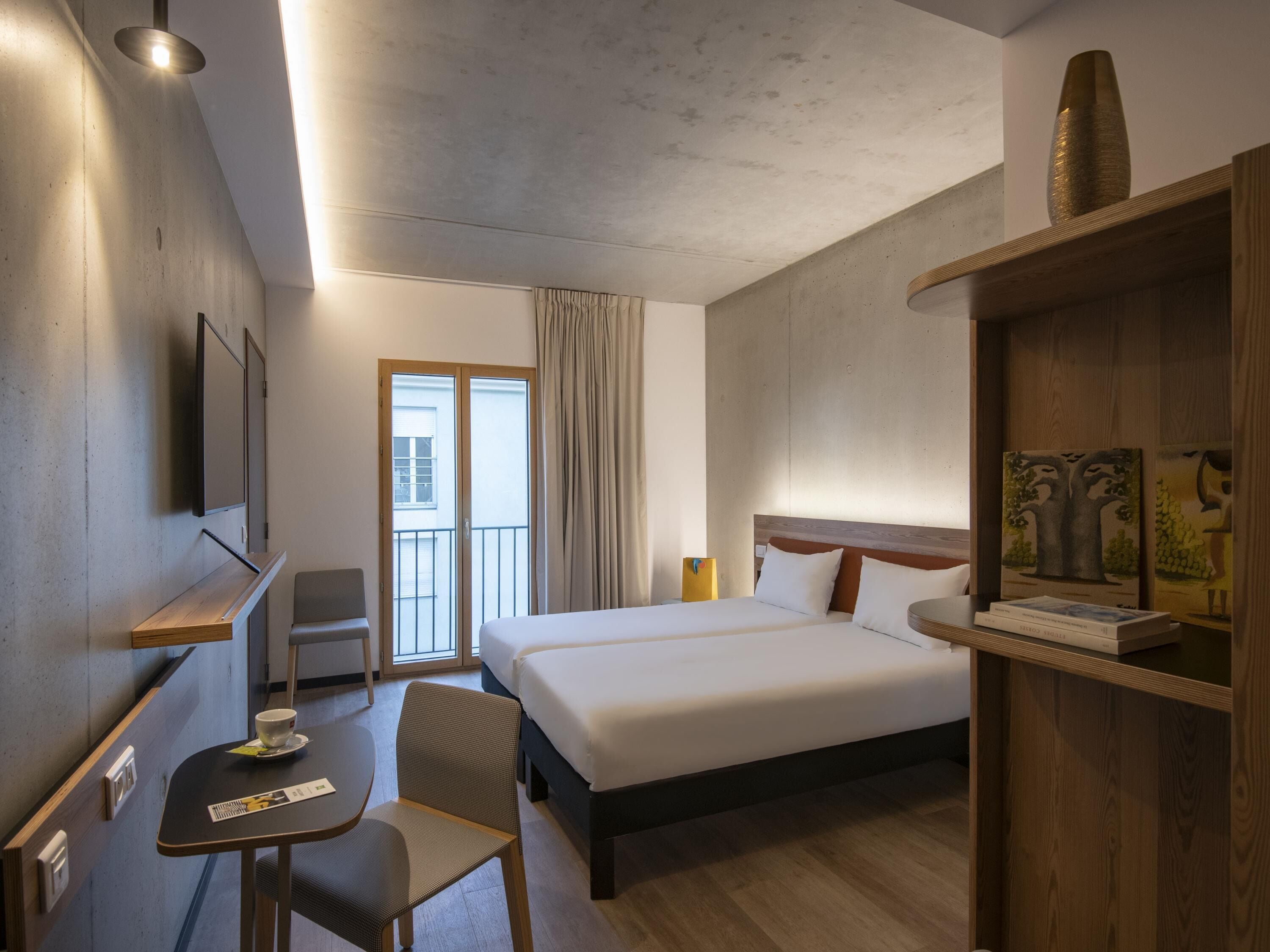 A hotel room with a bed, a desk with a chair, a television mounted on the wall, and a window with curtains.