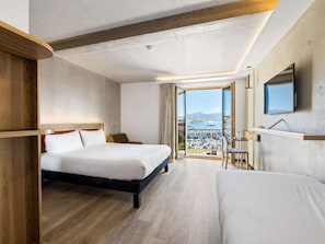 A hotel room with a bed, a desk, a chair, a TV, and a large window with a view of the city. - ibis Styles Ajaccio Napoleon (Ajaccio)