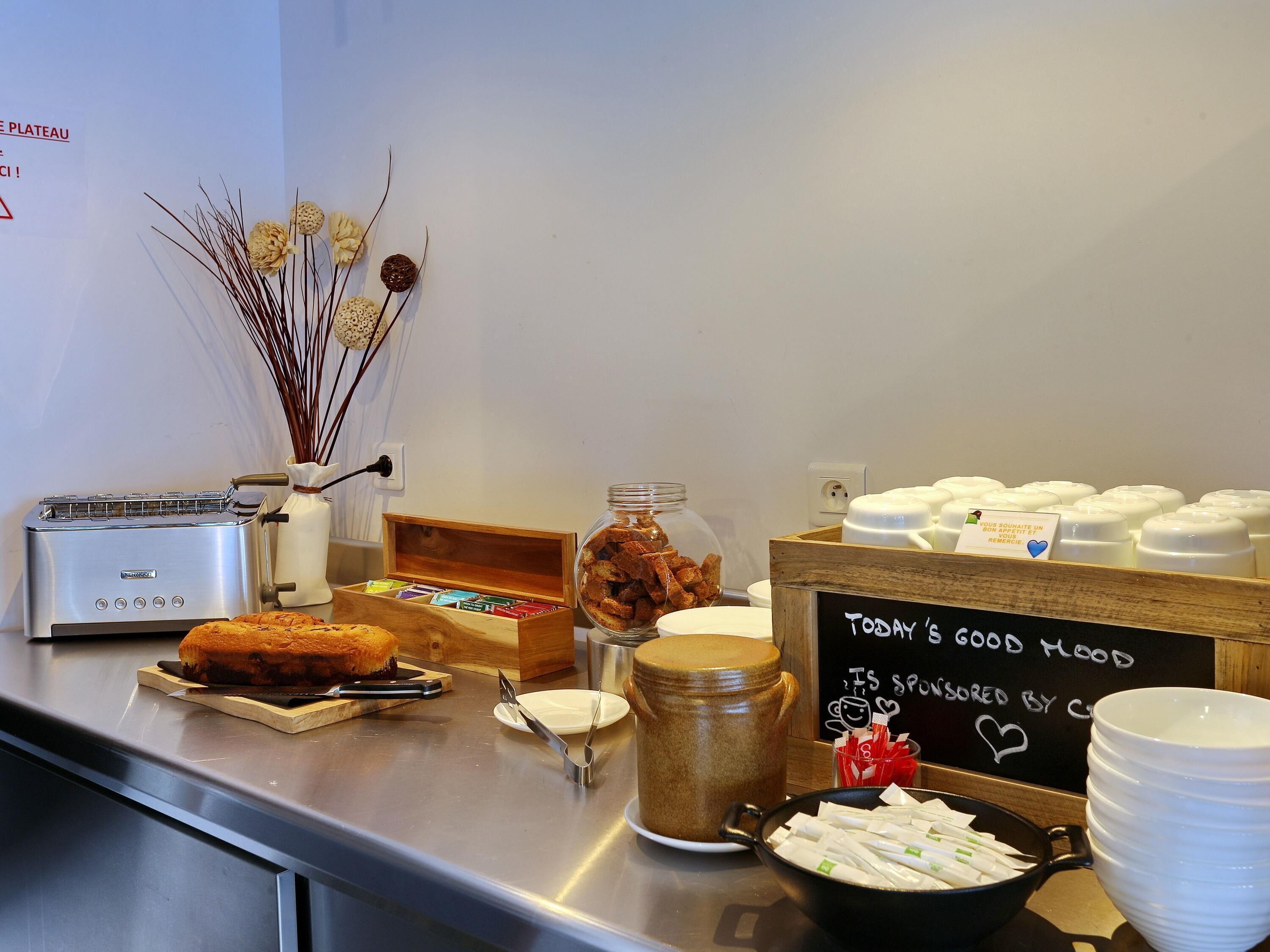 A breakfast buffet with a toaster, a wooden box of pastries, a jar of cereal, a coffee pot, and a sign reading 'Today's good mood'.
