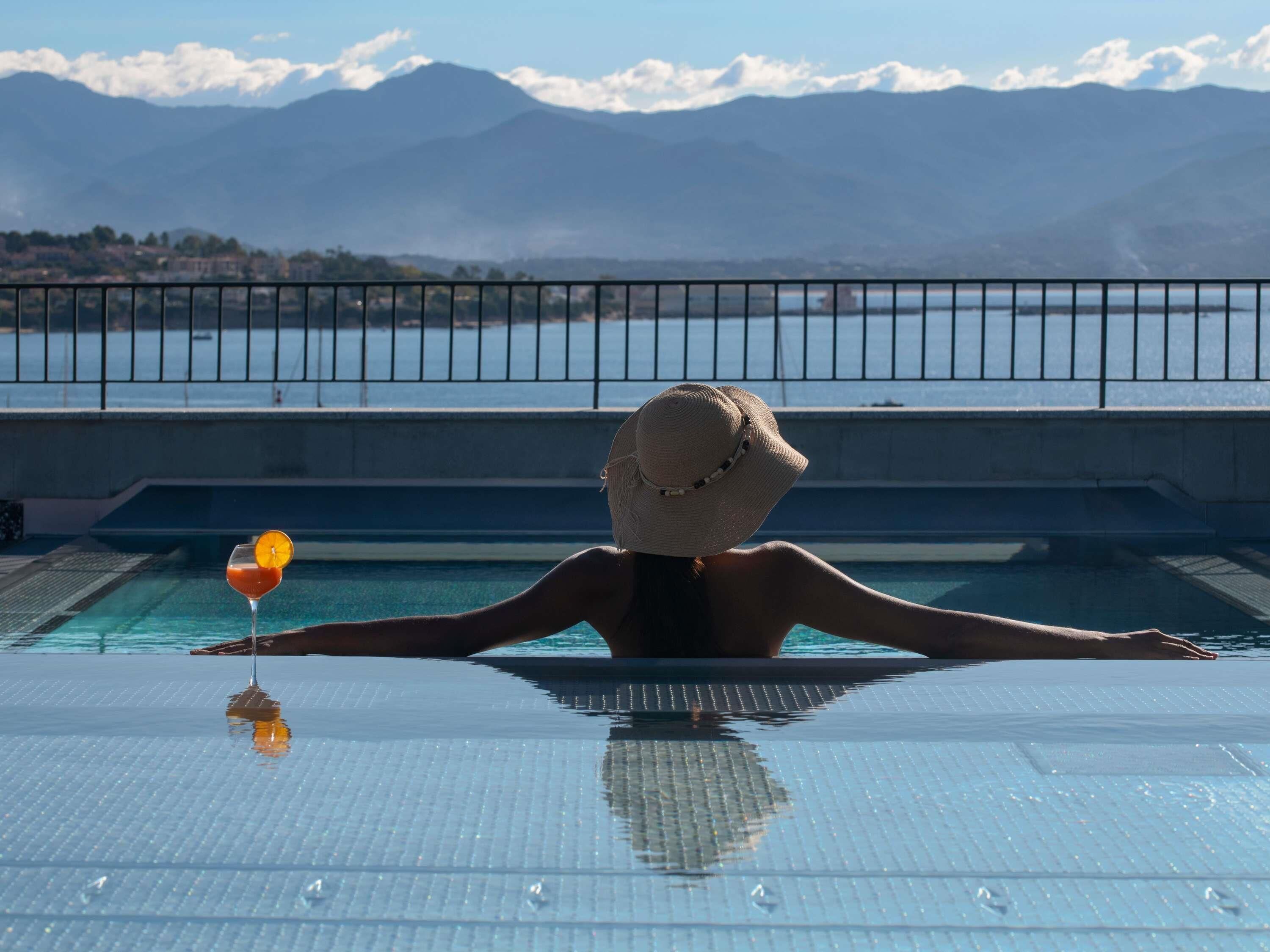 En person som kopplar av i en pool med en drink i handen och utsikt över bergen.