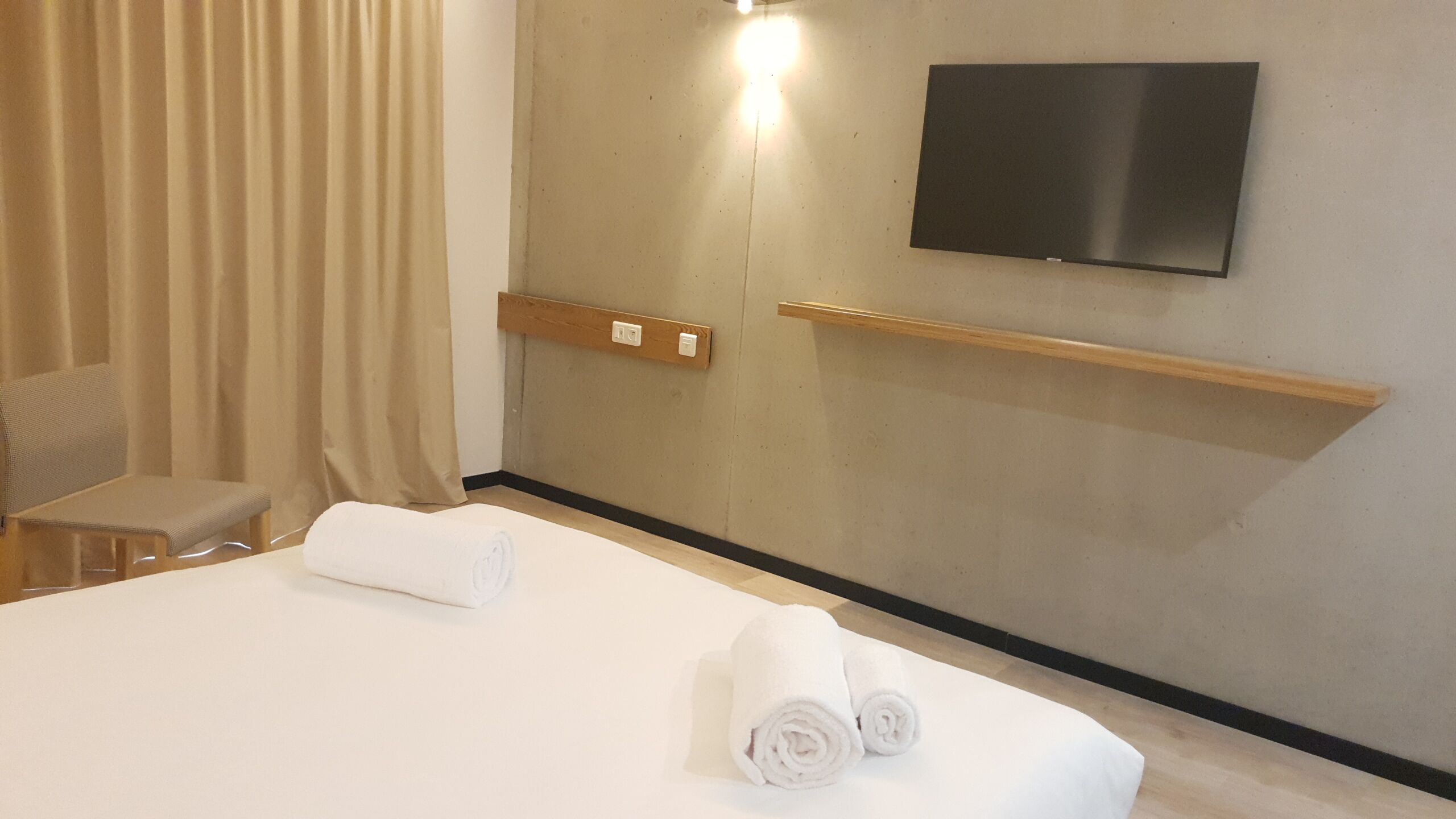 Una habitación de hotel con una cama, una silla, un televisor montado en la pared y un estante de madera.