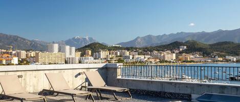 A rooftop pool with lounge chairs, a view of a cityscape, and mountains in the distance.