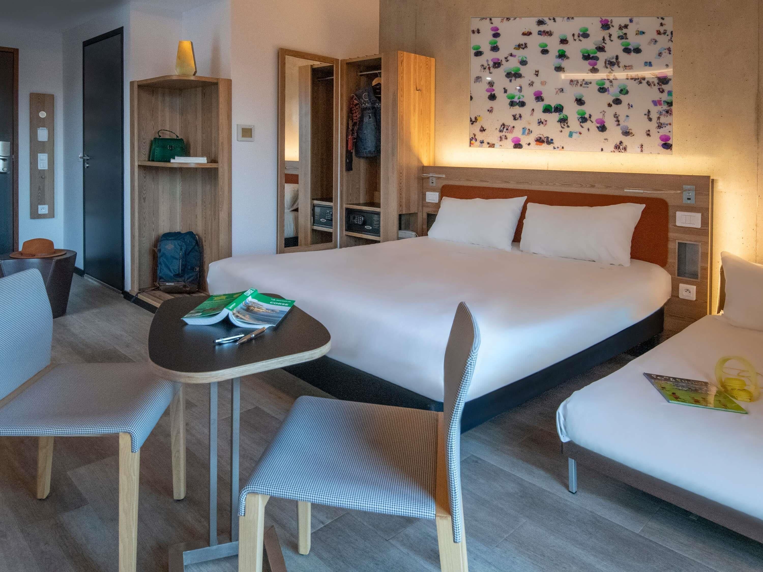 A hotel room with a bed, two chairs, a small table, a wardrobe, and a wall with a patterned artwork.