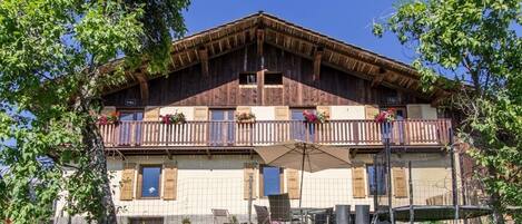 A two-story wooden chalet with balconies, outdoor seating, and a stone wall in front.