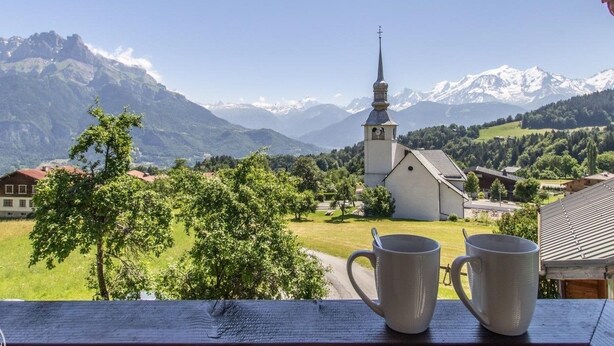 Vista de un paisaje montañoso con una iglesia y casas, y dos tazas blancas sobre una mesa de madera.