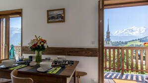 Un comedor con una mesa dispuesta para dos, un jarrón de flores y vistas a montañas a través de amplias ventanas.