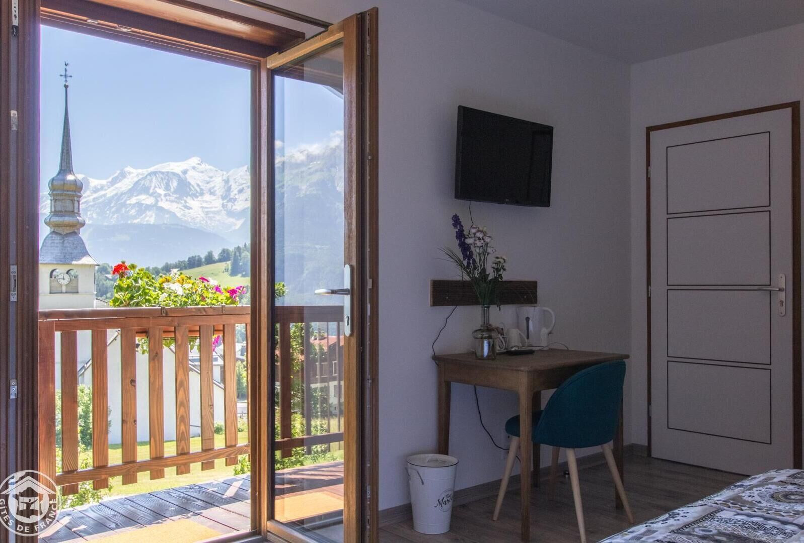 A room with a balcony, a desk with a chair, a TV mounted on the wall, and a door.