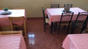 A dining area with two tables, each covered with a pink tablecloth. There are wooden chairs around the tables, and a potted plant is placed on one table. The floor is tiled with a reddish-brown color. - Semo Hotel (Gumare)