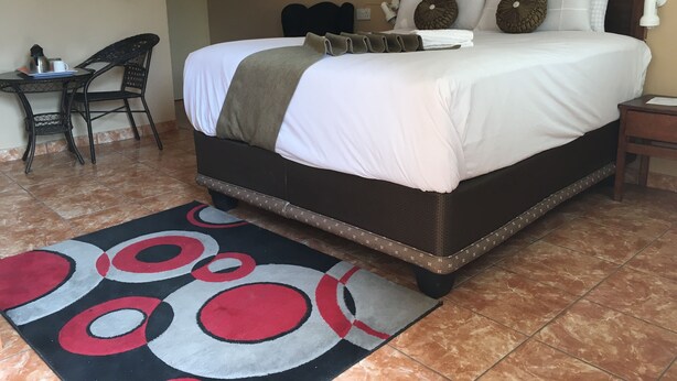 A neatly made bed with a white comforter and pillows, a brown headboard, a bedside table with a lamp, and a patterned rug on a tiled floor. - Semo Hotel (Gumare)