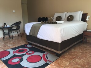 Una cama bien hecha con edredón blanco y almohadas, cabecera marrón, mesita de noche con lámpara y alfombra estampada sobre piso de baldosas.