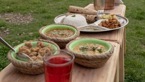 Una mesa de madera con diversos platos y una jarra de metal, al aire libre con un edificio pequeño al fondo.