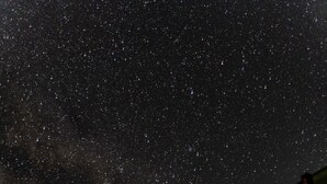 Cielo nocturno lleno de estrellas, un edificio con techo inclinado y silueta de árboles.