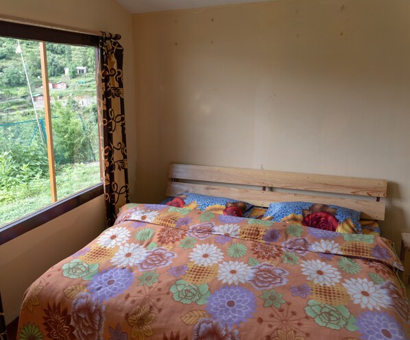 A bedroom with a wooden headboard, a floral bedspread, and a window with a view of greenery outside. - The Goat Village Escape (Nainital)