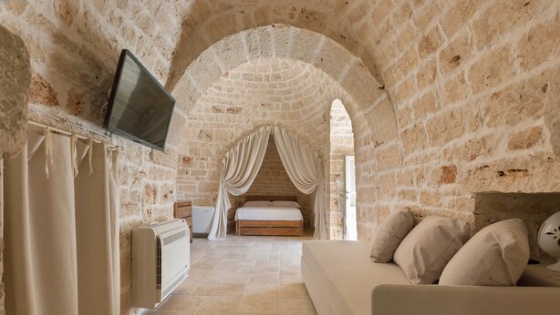 A room with a stone wall, a bed with a canopy, a TV mounted on the wall, and a sofa. - Trulli di Buontempo (Francavilla Fontana)