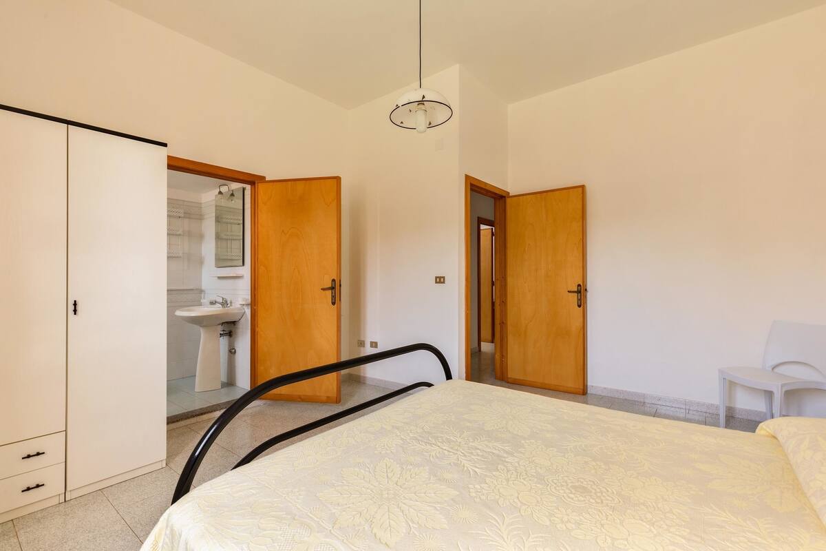 Ein Schlafzimmer mit Bett, Kleiderschrank, Stuhl und einem durchgehenden Türrahmen sichtbarem Badezimmer.