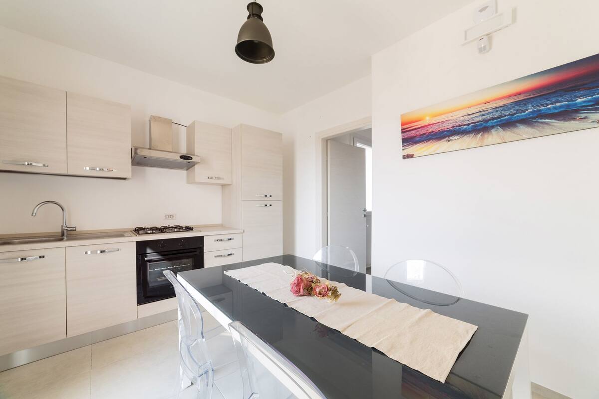 Uma cozinha moderna com armários de madeira clara, bancada preta e mesa de jantar de vidro transparente.