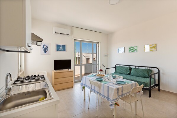A compact kitchen with a gas stove, sink, and a television. A dining table with chairs is set up in the center, and a green sofa is against the wall.