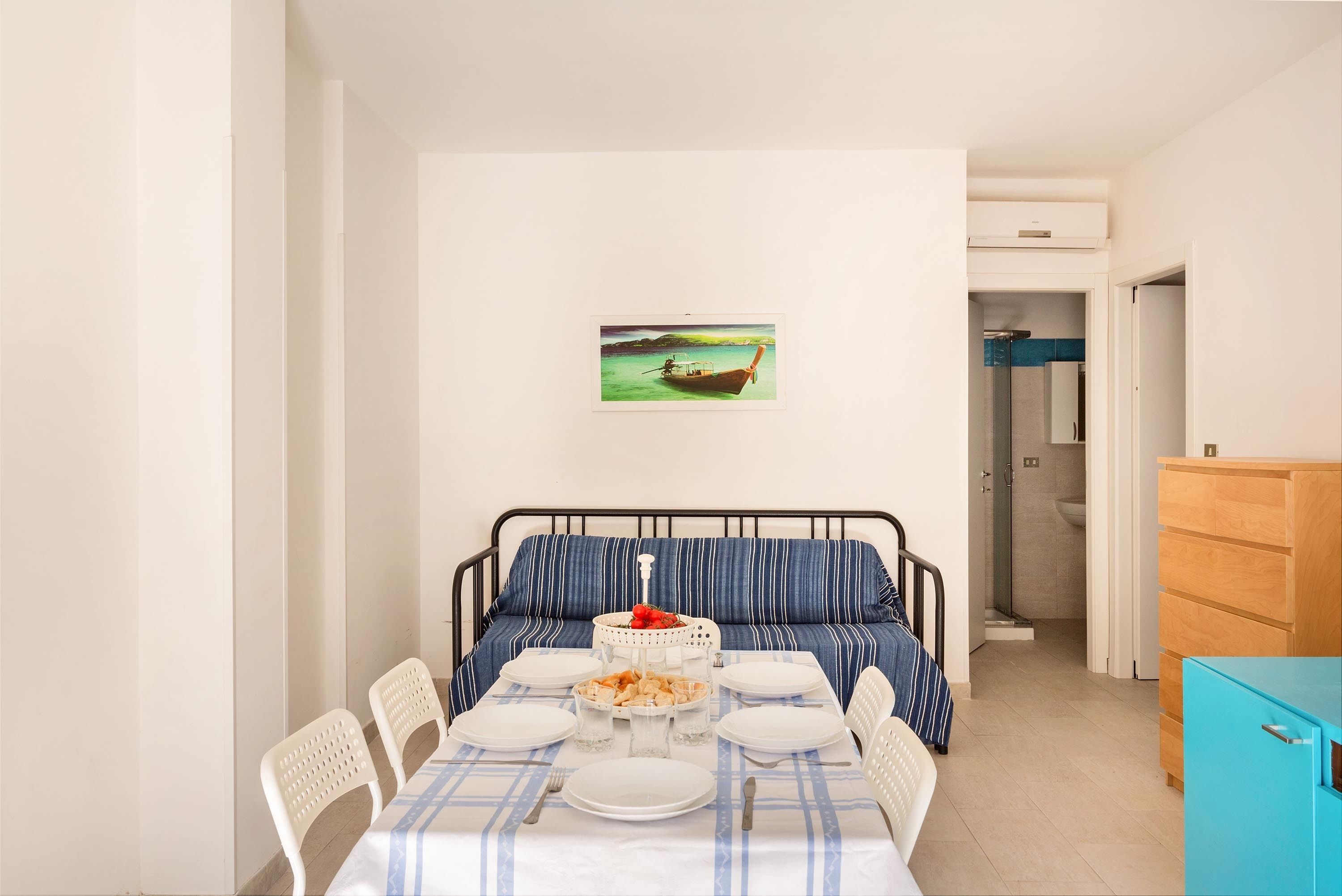 A neatly arranged dining area with a table set for four, a bed with striped bedding, a small painting on the wall, and a view into the bathroom.