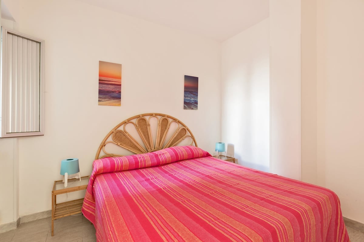 Une chambre à coucher avec un lit, deux tableaux accrochés au mur, une fenêtre et une table de chevet avec une lampe.
