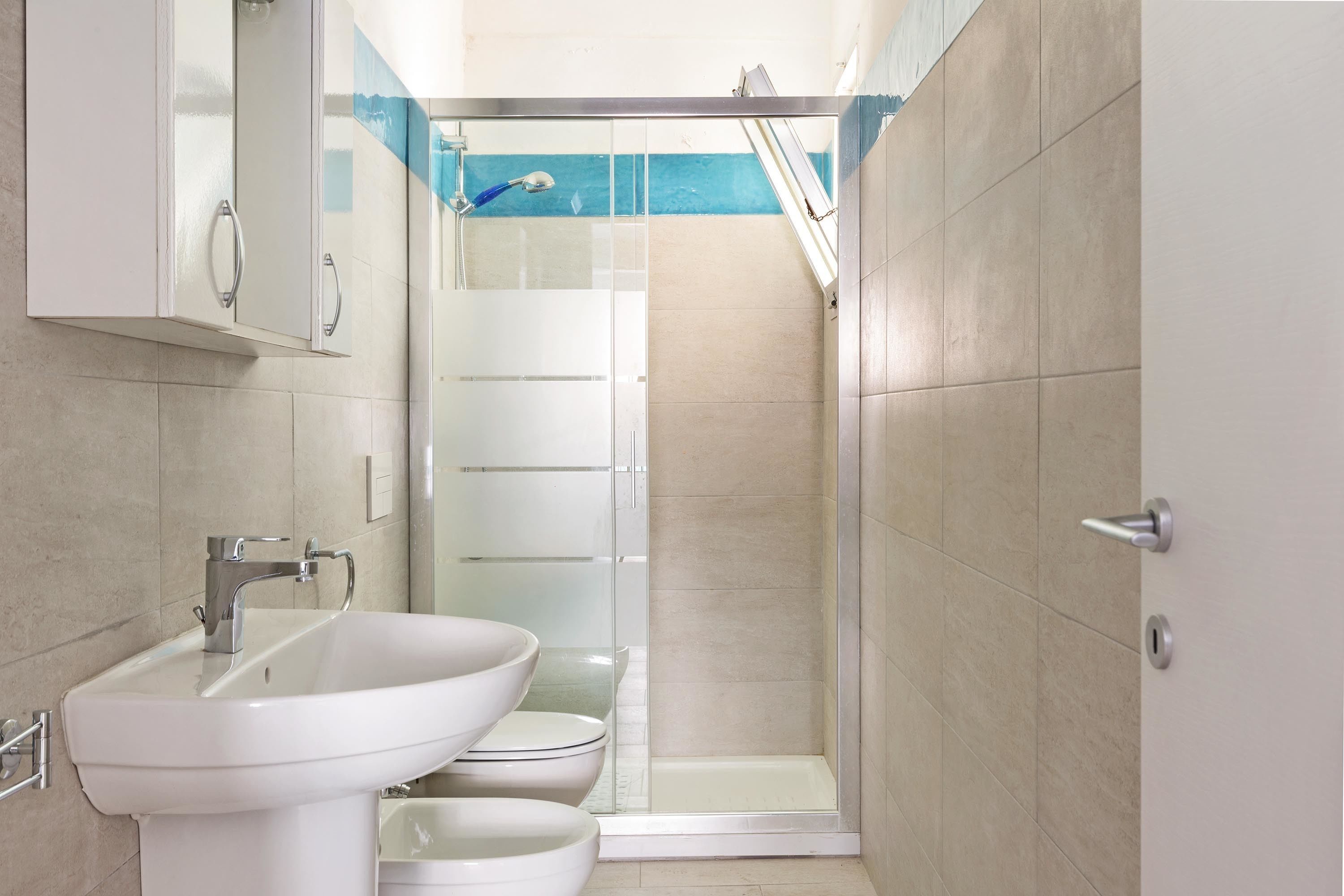 A modern bathroom with a white sink, two toilets, a glass shower enclosure, and a door with a silver handle.