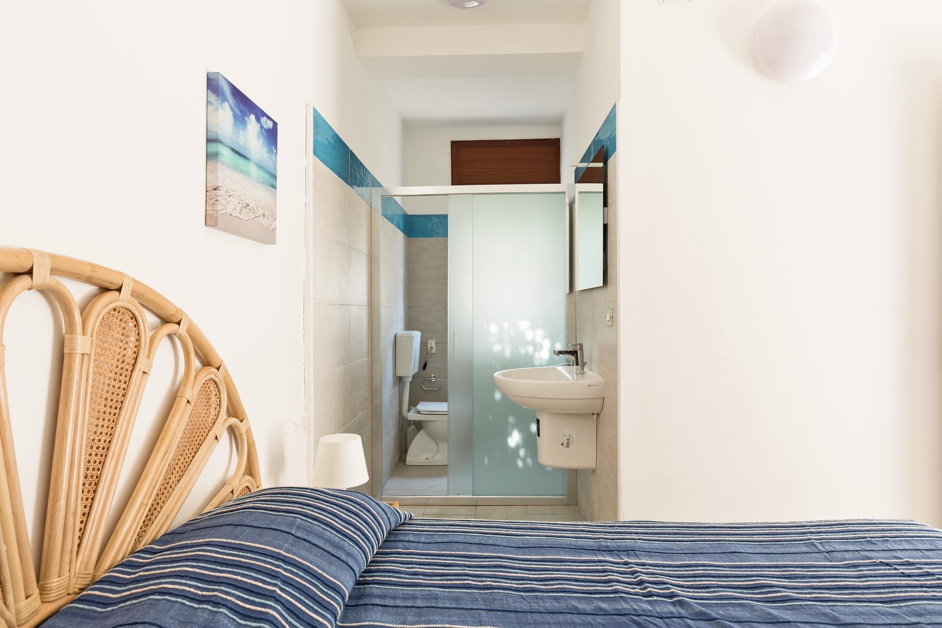 A bedroom with a bed, a rattan headboard, a striped pillow, a bathroom with a glass shower enclosure, a sink, and a mirror.