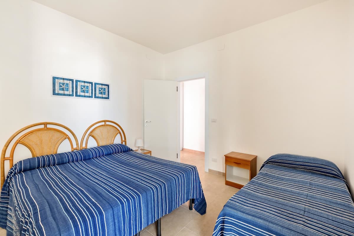 Une chambre avec deux lits, une tête de lit en bois, une petite table de chevet avec une lampe et une porte donnant sur une autre pièce.