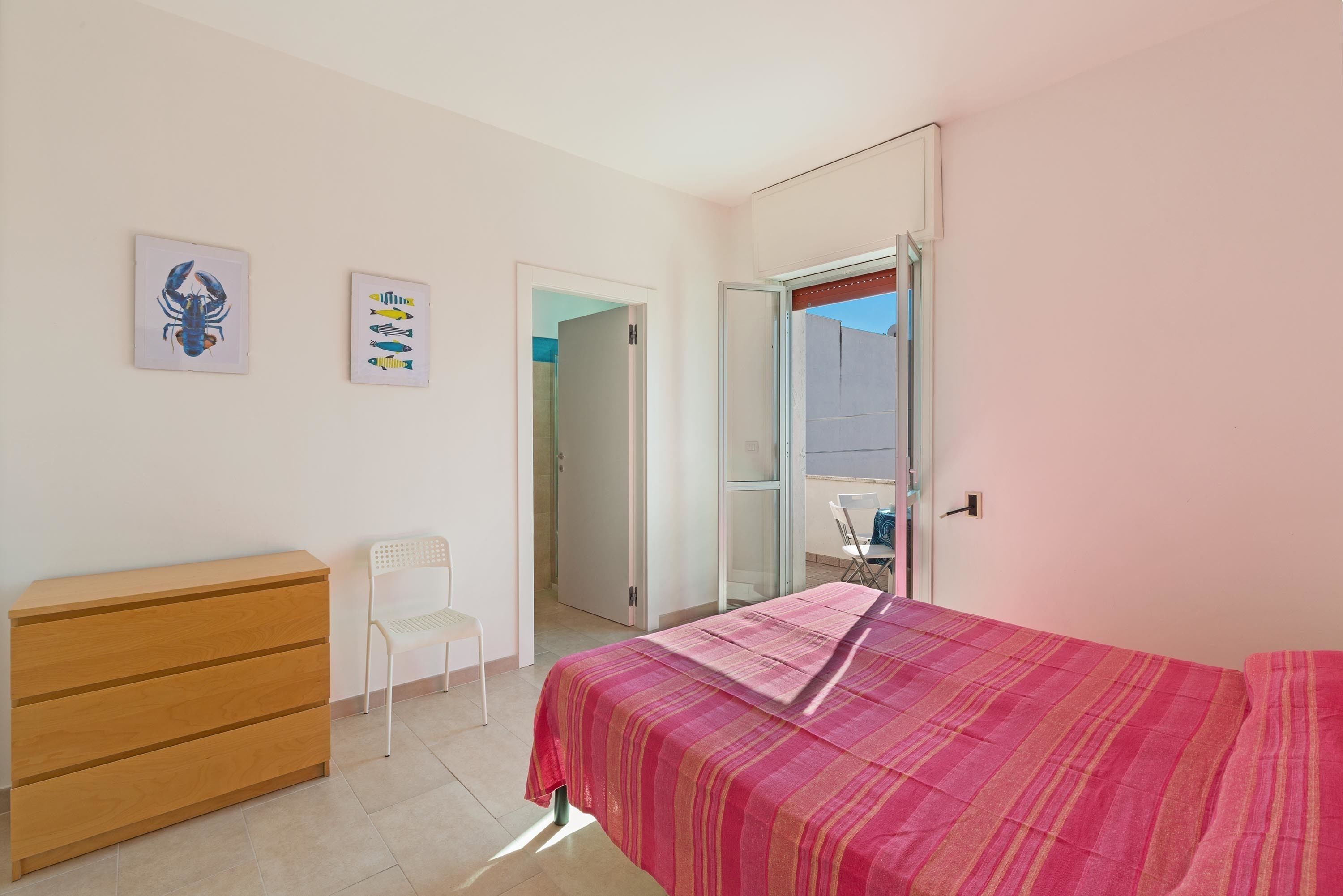Un dormitorio con una cama, un aparador de madera, una silla blanca y dos cuadros colgados.