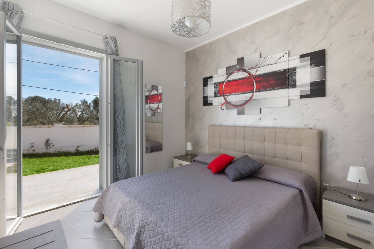 Une chambre à coucher moderne avec un grand lit, une porte vitrée donnant sur un balcon et une œuvre d’art contemporaine accrochée au mur.