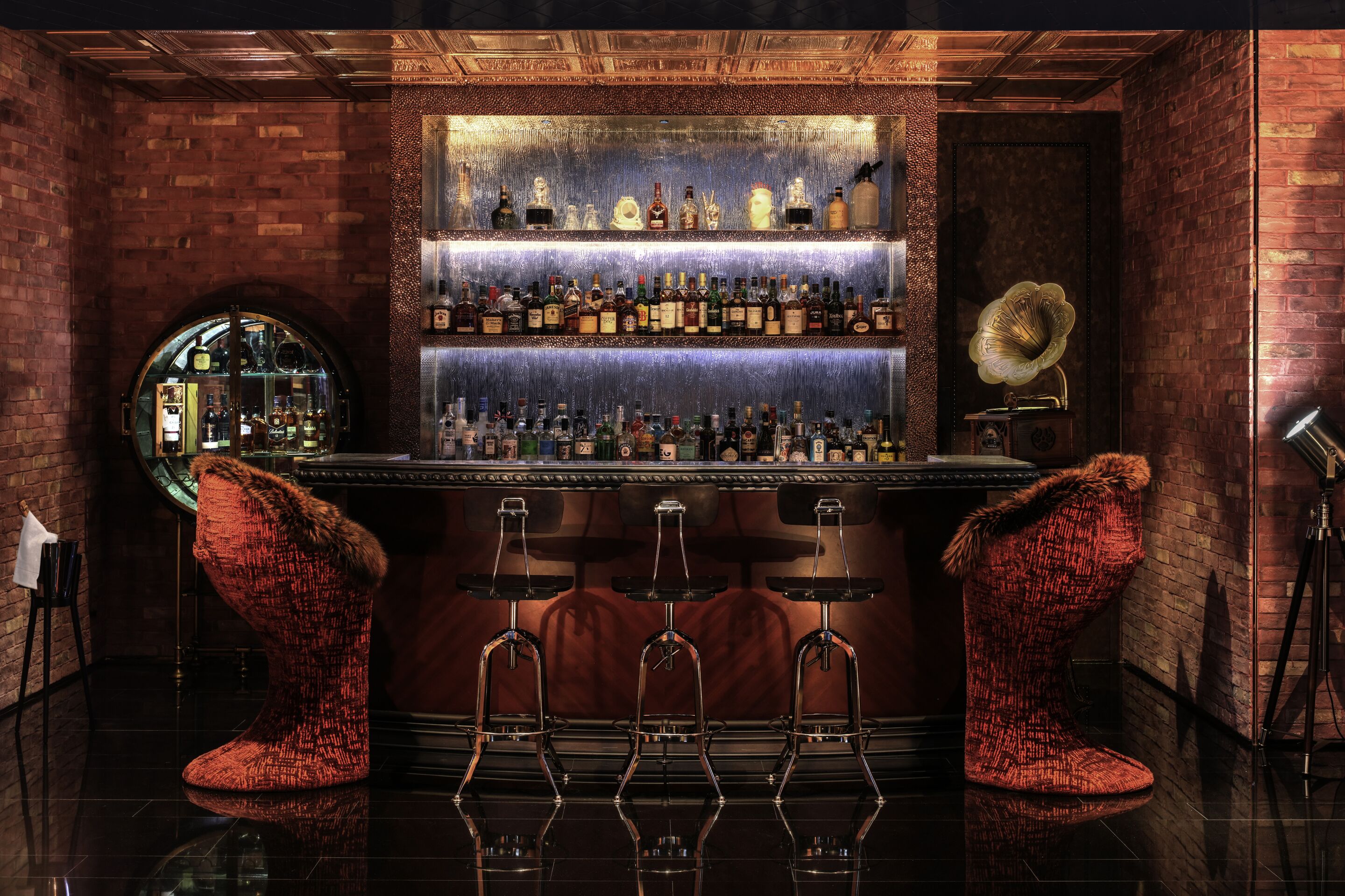 A bar with a brick wall, shelves of bottles, and two red upholstered stools.
