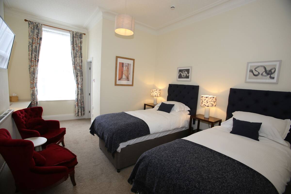 A hotel room with two beds, a red armchair, a small table, and a window with curtains.