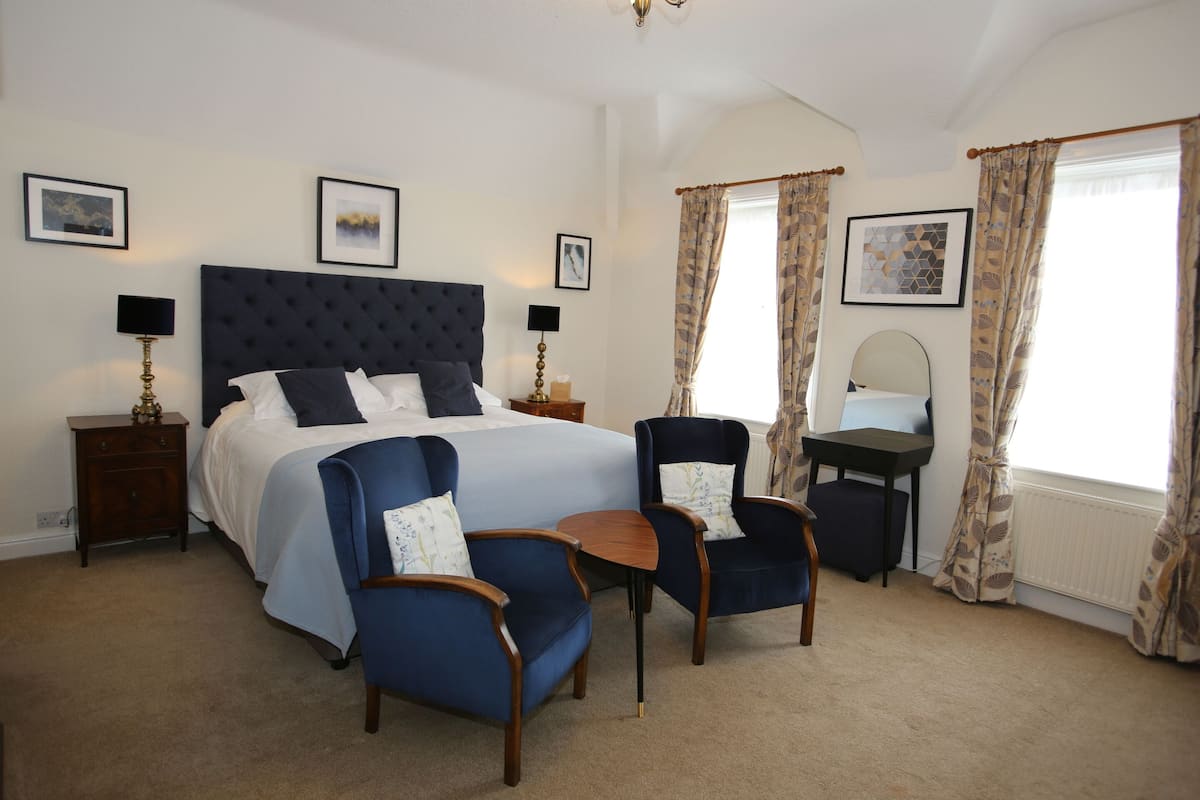 A bedroom with a bed, two armchairs, a small table, a nightstand, a desk, and a mirror.