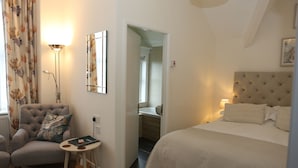 A bedroom with a bed, a chair, a small table, a lamp, a mirror, and a bathroom visible through an open door. - The Cordelia (Scarborough)