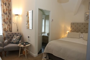 A bedroom with a bed, a chair, a small table, a lamp, a mirror, and a bathroom visible through an open door. - The Cordelia (Scarborough)